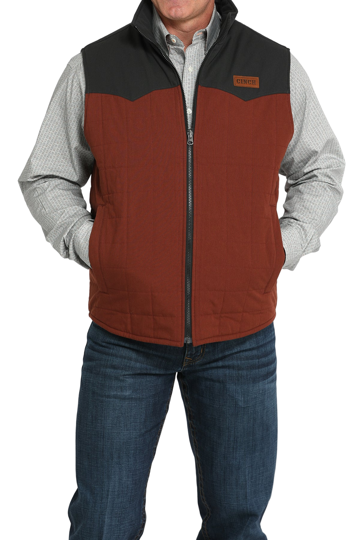 A man wears a two-tone orange and brown zip-front vest over a light gray checkered shirt and jeans