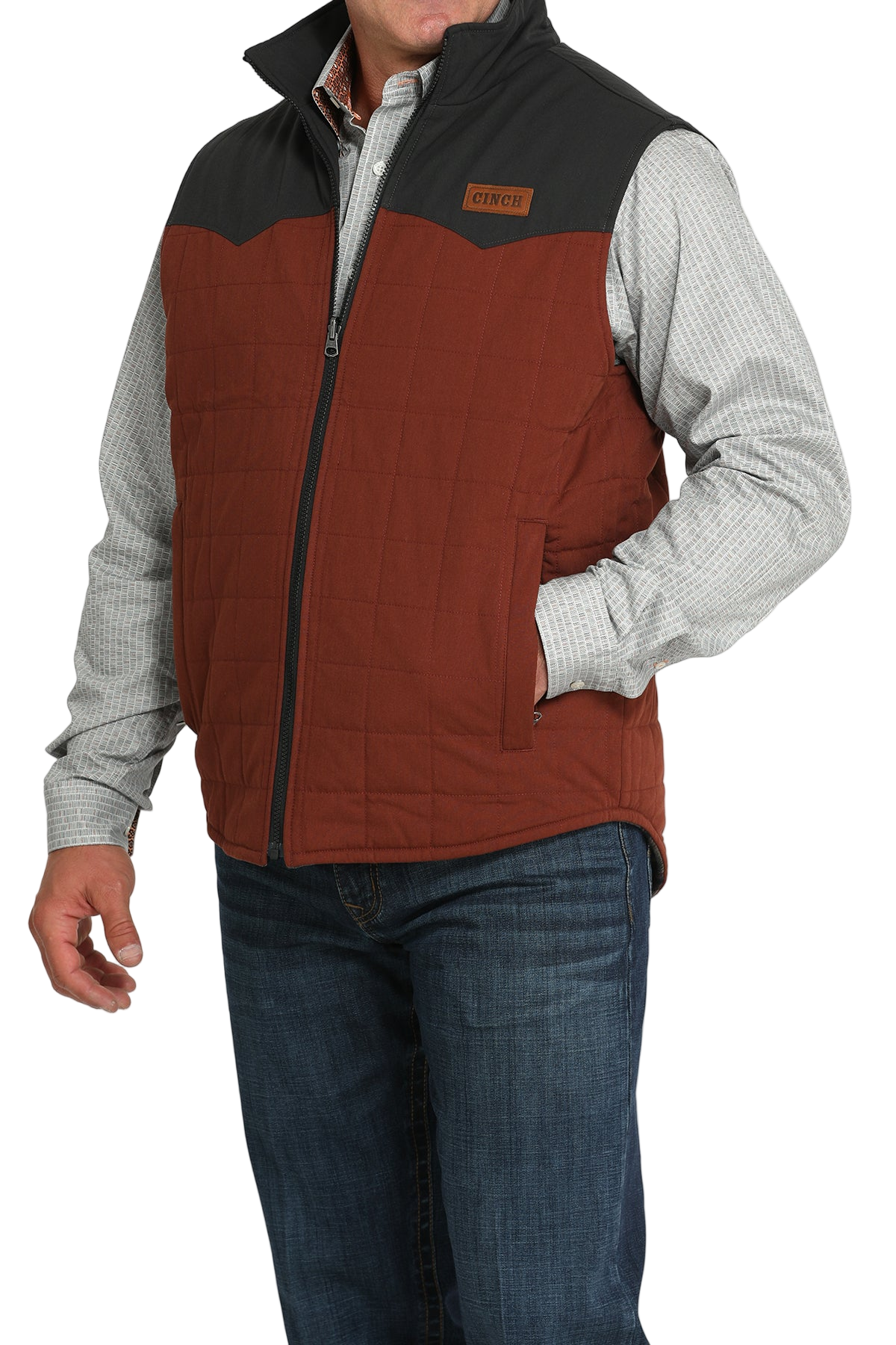 A man wears a two-tone orange and brown zip-front vest over a light gray checkered shirt and jeans