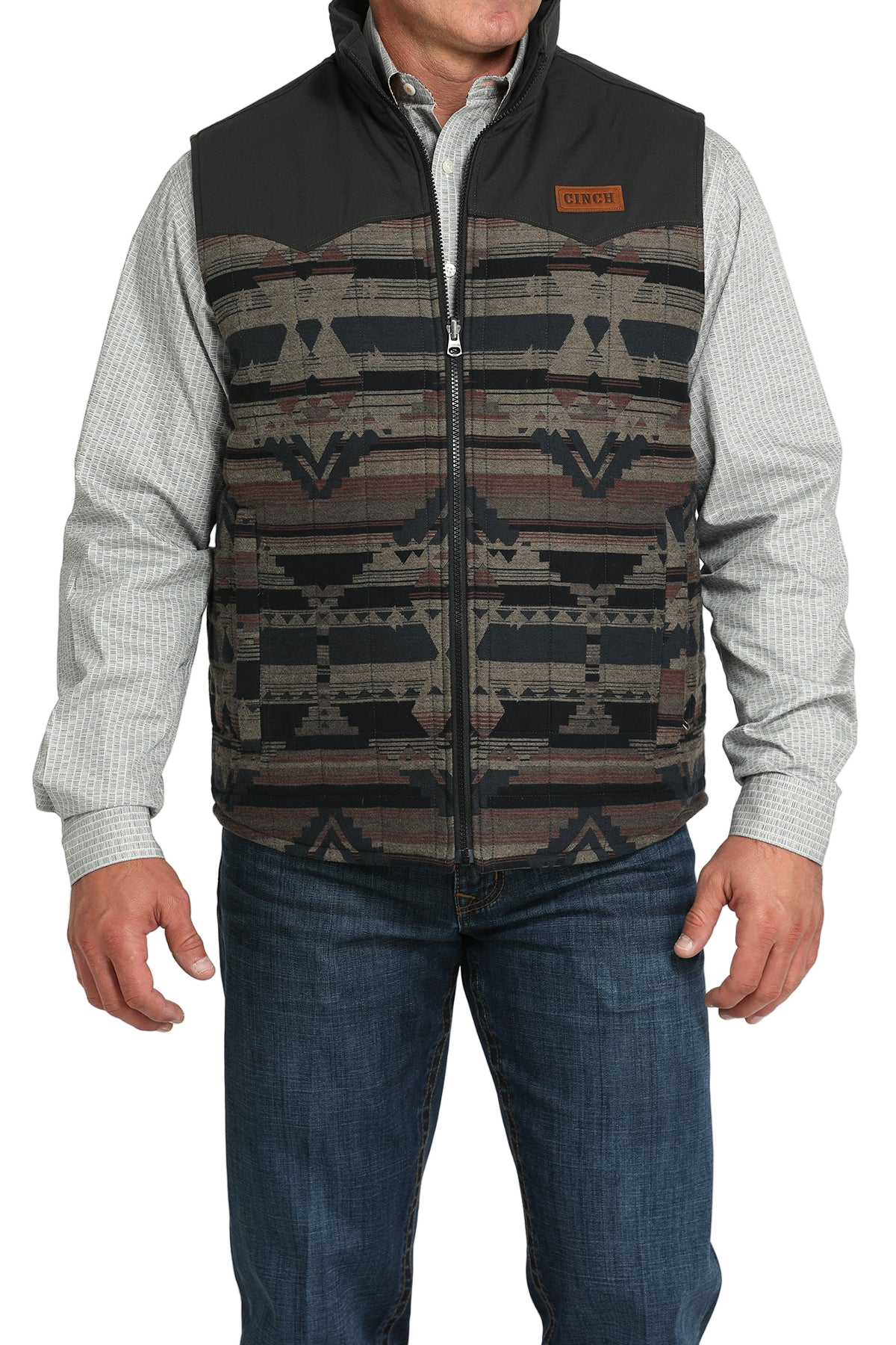 A man wears a patterned zip-front vest with black shoulders over a light gray checkered shirt and jeans