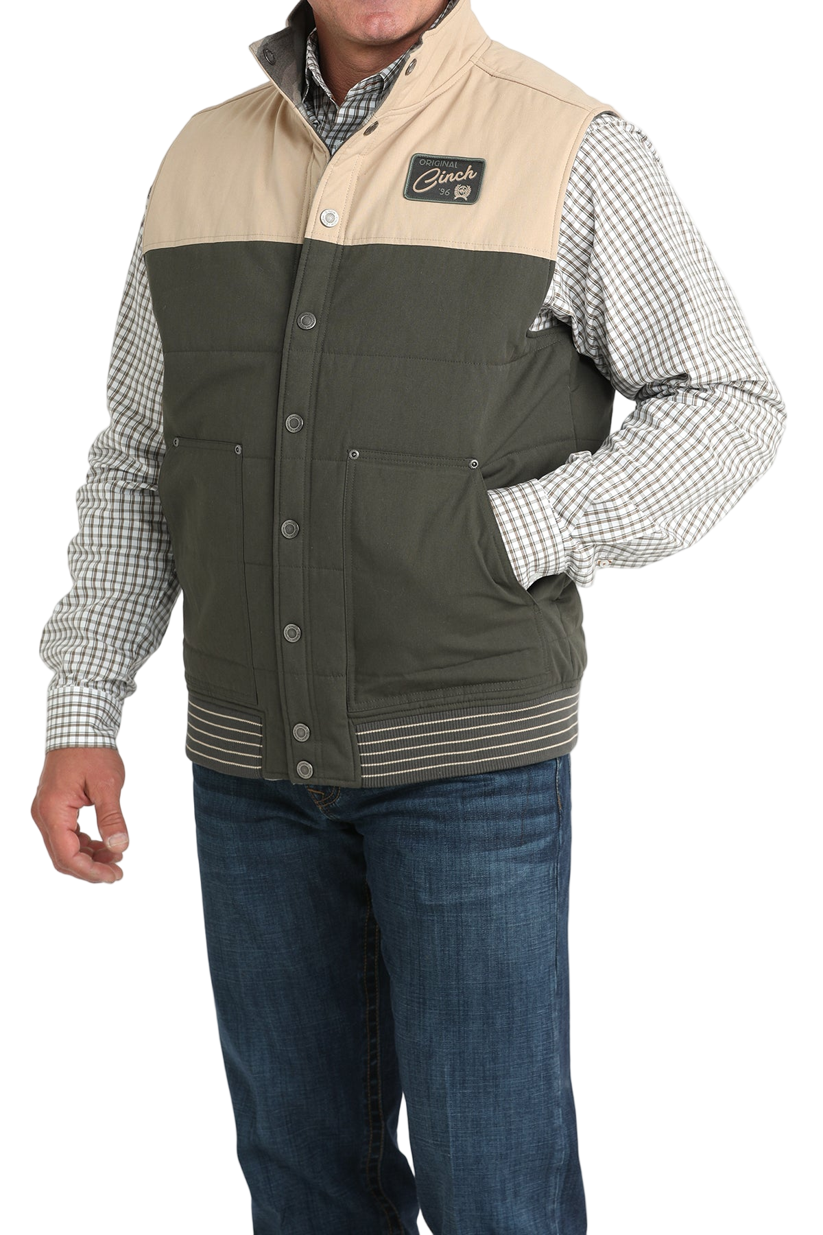 A man wearing a two-tone vest with a beige top panel and dark green lower panel over a checkered button-down shirt, paired with blue jeans