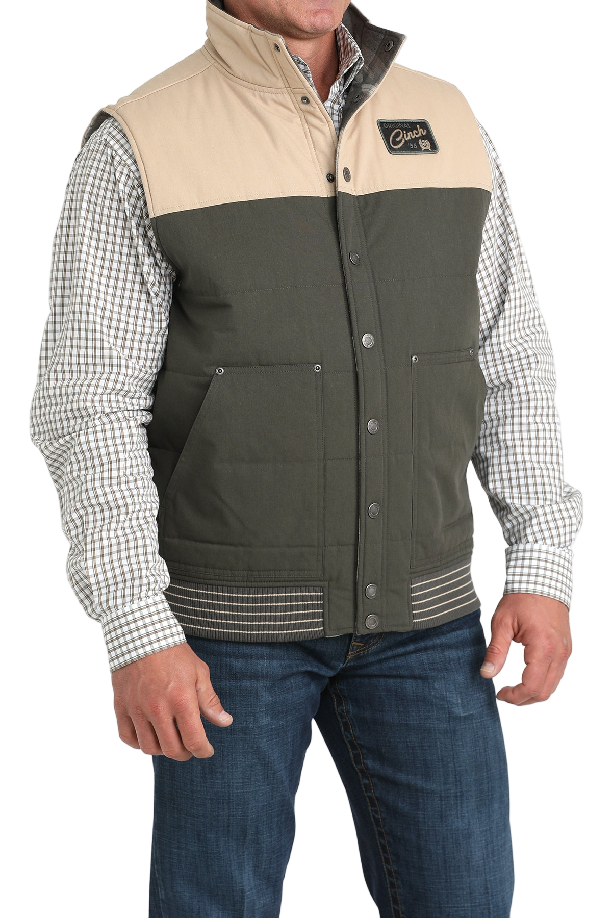 A man wearing a two-tone vest with a beige top panel and dark green lower panel over a checkered button-down shirt, paired with blue jeans