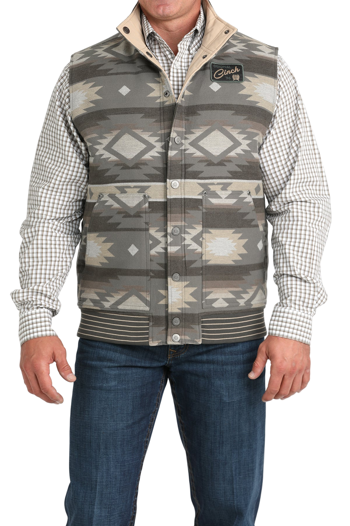 A man wearing a gray and beige Aztec-patterned vest over a checkered button-down shirt, paired with blue jeans