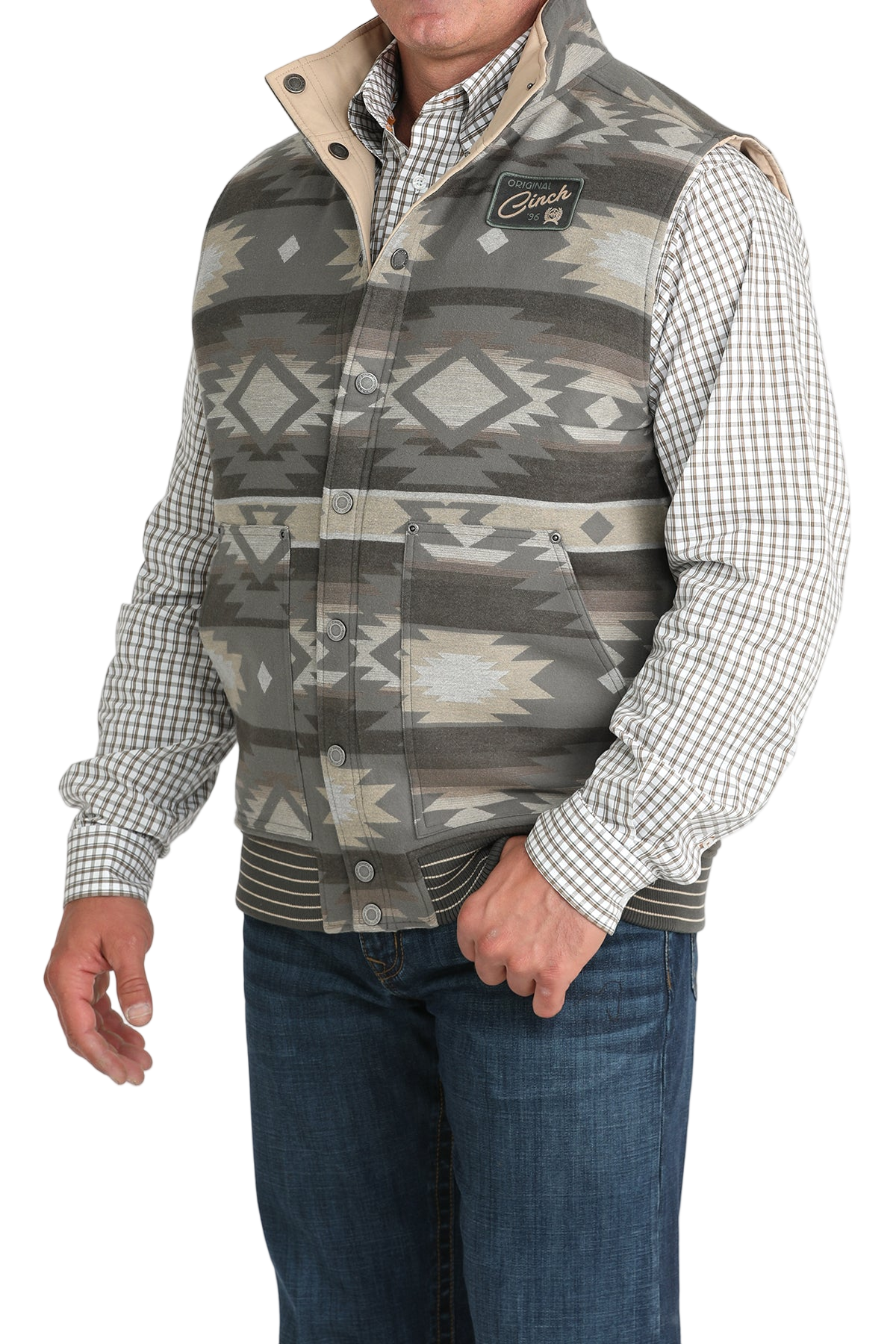 A man wearing a gray and beige Aztec-patterned vest over a checkered button-down shirt, paired with blue jeans