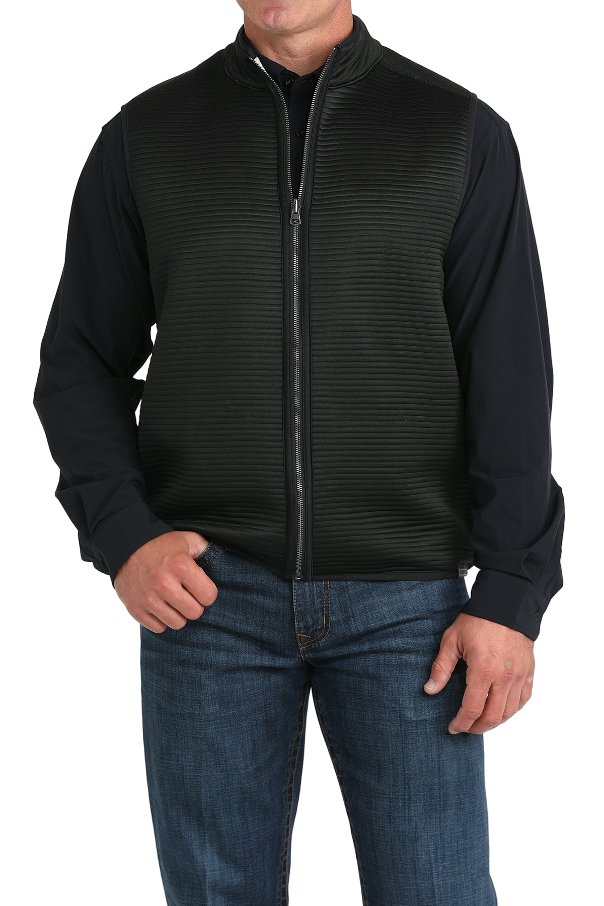 A man wears a black zip-front vest over a black long-sleeve shirt and blue jeans