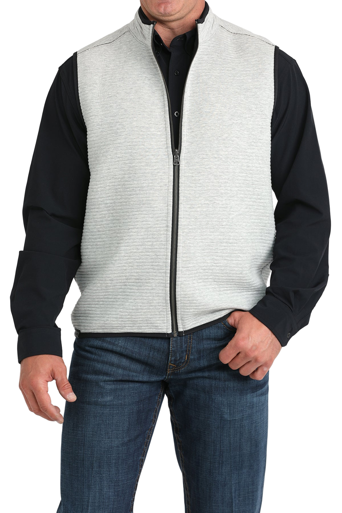 A man wears a light gray zip-front vest over a black long-sleeve shirt and blue jeans