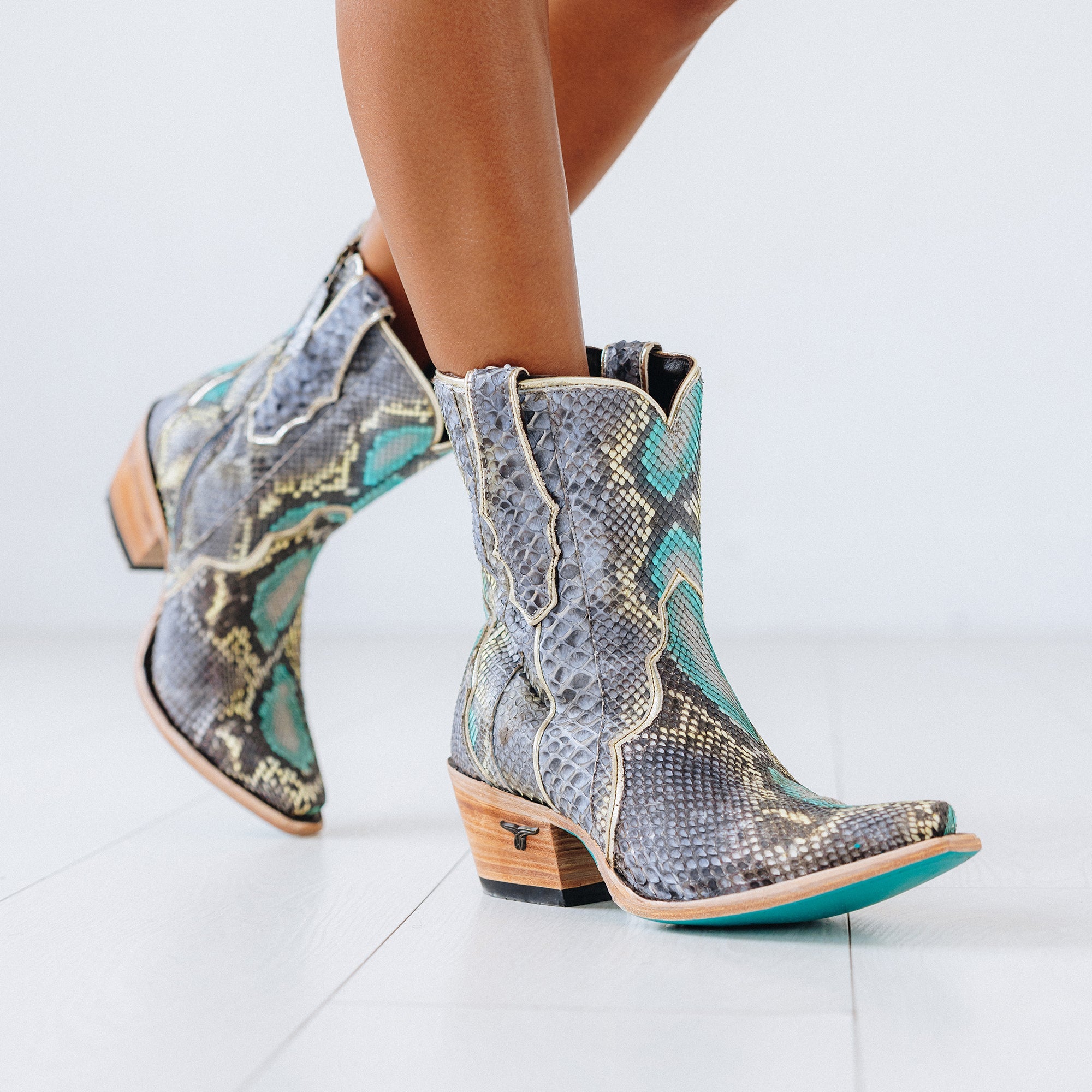 Pair of short cowboy boots with a bold snakeskin pattern in shades of gray, turquoise, and cream. The boots feature a snip toe, stacked wooden heel, and turquoise soles for a striking contrast