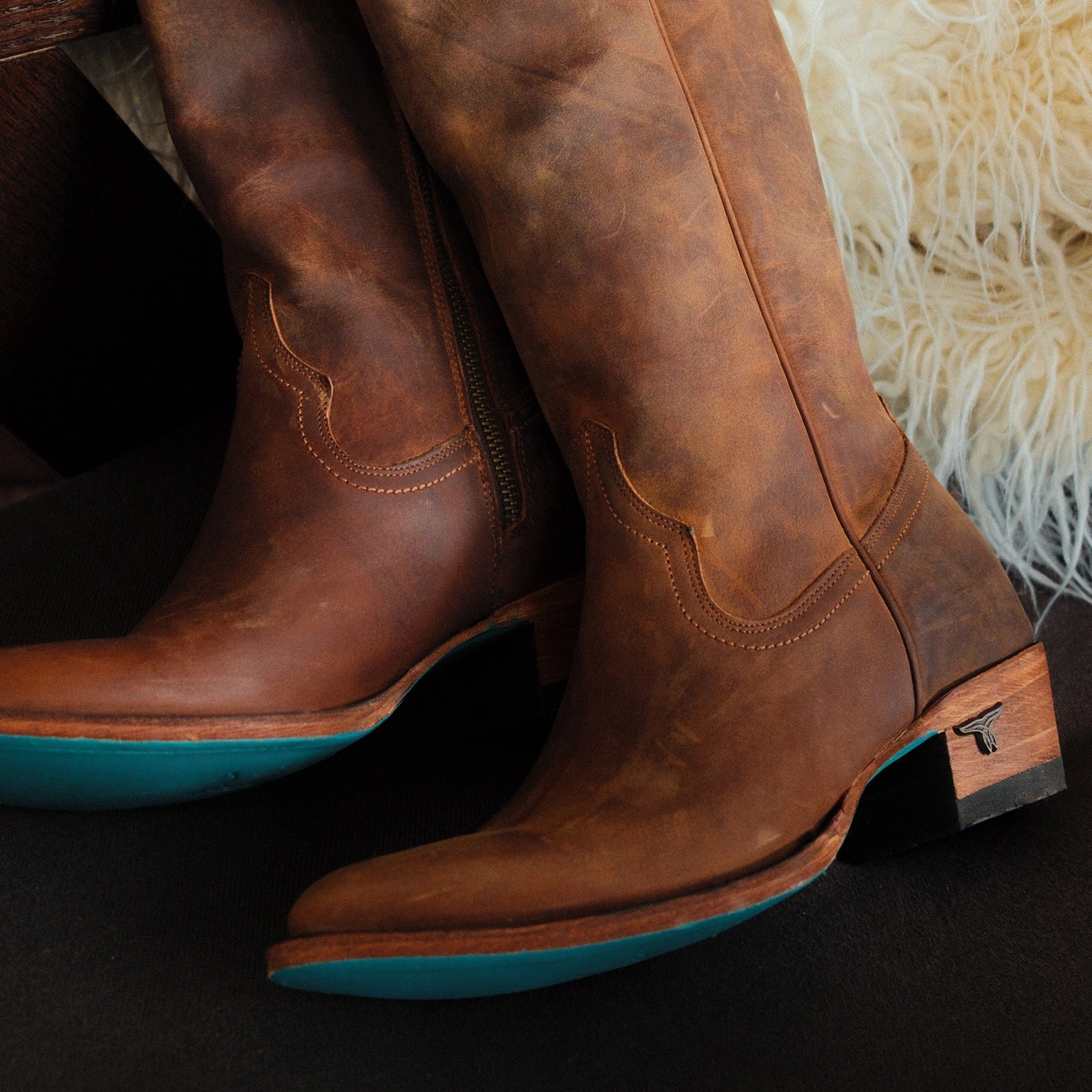 Pair of brown cowboy boots with subtle stitching, wooden heels, and turquoise soles resting on a dark surface next to a fluffy cream throw