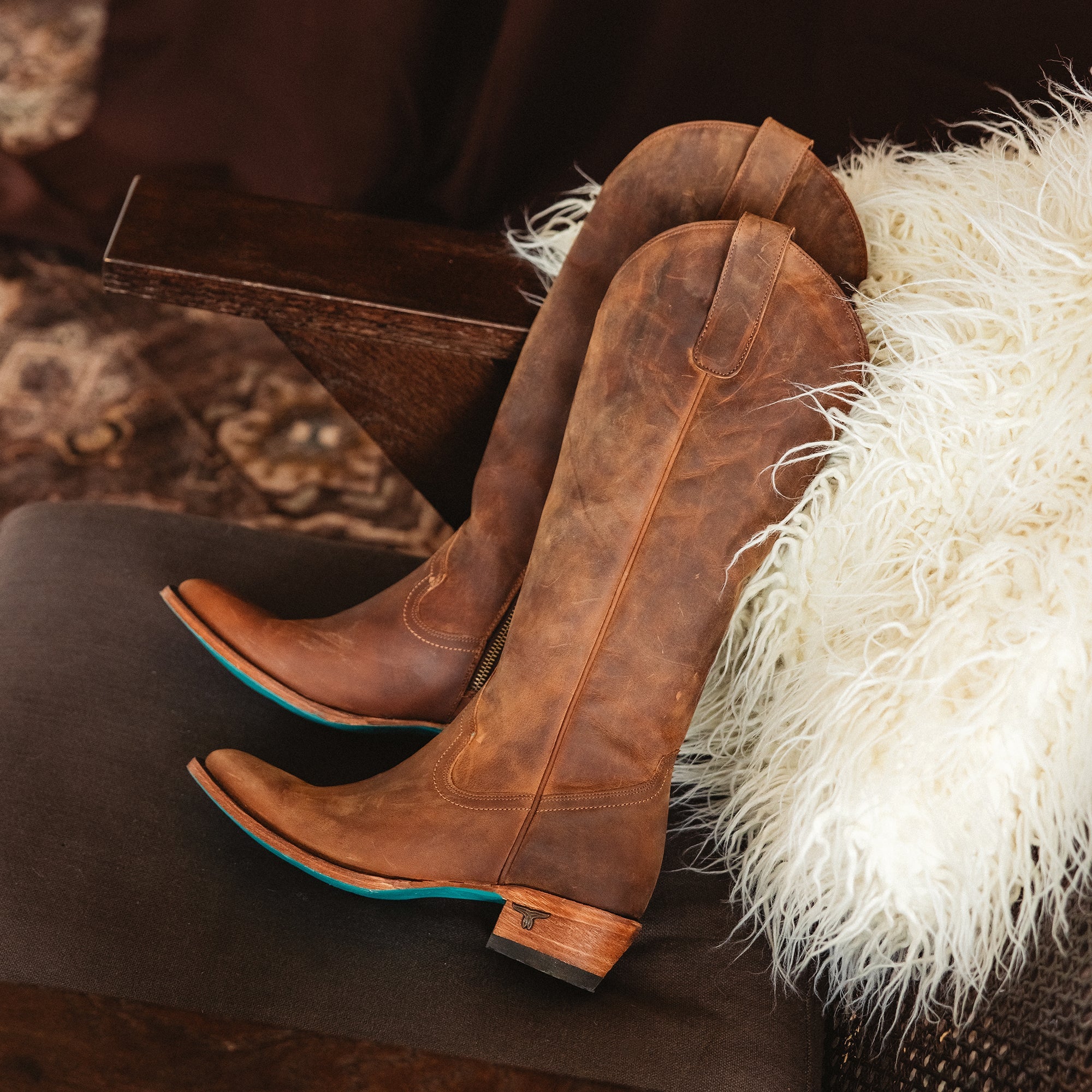 Pair of brown cowboy boots with simple stitching, wooden heels, and turquoise soles resting on a chair with a fluffy cream throw