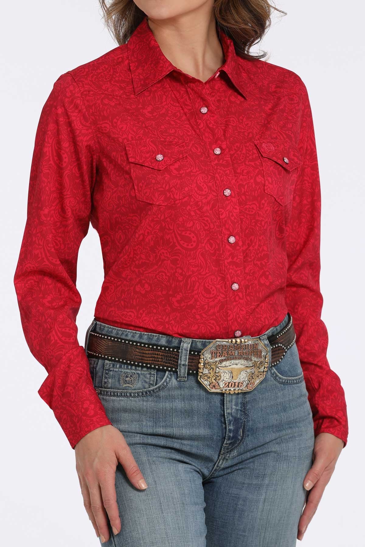 Woman wearing a pink long-sleeve button-down shirt with subtle paisley pattern and western-style chest pockets, paired with blue jeans and a western belt buckle displayed on a white background.